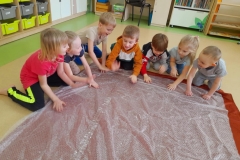 Grupa dzieci siedzi wokół folii bąbelkowej, uderzają dłońmi we folię w rytm muzyki