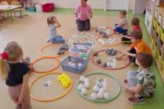 Kilkoro dzieci siedzi na podłodze, przed nimi obręcze, w których są małe piłeczki i ułożone działania matematyczne.