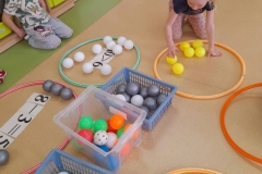 Dziewczynka liczy żółte piłeczki w obręczy, w dwóch pojemnikach leżą kolorowe piłeczki.