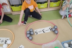 Chłopiec siedzi na podłodze, przed nim obręcz w której są małe piłeczki. Chłopiec układa działanie matematyczne.