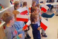 Grupa dzieci stoi wraz z wychowawczynią, w ręku trzymają flagi Polski, śpiewają pieśń Przybyli ułani.