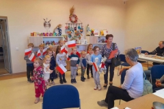 Grupa dzieci stoi, w rękach trzymają flagi Polski, wychowawczyni odczytuje życzenia z laurki dla seniorów.