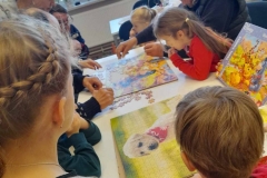 Seniorzy układają na stole puzzle, wokół stołu dzieci, pomagają w układaniu puzzli.