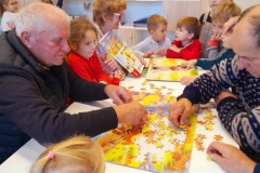 Seniorzy układają puzzle na stole, wokół stołu przedszkolaki, pomagają układać puzzle.