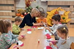 pięcioro dzieci siedzi przy stoiku wraz z panią. każde ma nożyczki  w rękach. Na stoliku leża białe i czerwone serca.