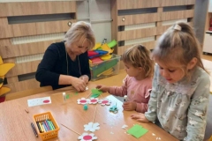 Dwie dziewczynki siedzą przy stoliku. Pani kuca obok nich. Na stoliku leża białe okrągłe kwiatki z czerwonymi oczkami.