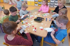 Sześcioro dzieci siedzi przy stoliku a obok kuca ani. Dzieci wycinają kwiatki.
