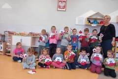 Grupa dzieci siedzi na podłodze. Za nimi stoją pozostałe wraz z panią. Zdjęcie wykonane na tle ściany.