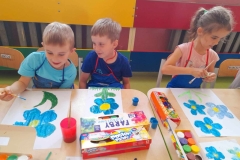 Dwóch chłopców I dziewczynka malują Niezapominajki. Na stole leżą farby.