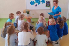 Grupa dzieci siedzi przed tablicą na której widać Niezapominajki, na stołku we flakonie też są Niezapominajki, wychowawczyni czyta opowiadanie.