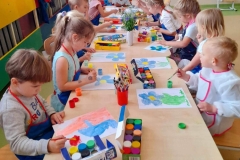 Dzieci siedzą po dwóch stronach stołu  malują farbami Niezapominajki. Na stole leżą farby, pędzle orz bukie żywych Niezapominajki.t