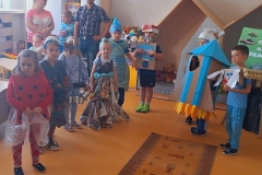 Grupa dzieci ubrana w stroje wykonane z różnych odpadów z lewej strony dywanika rozłożonego na podłodze . Z prawej strony stoi 2 chłopców jeden jako robot , drugi w przebraniu rakiety wykonanej z kartonowego pudełka.