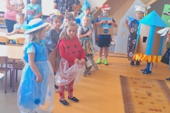 Grupa dzieci ubrana w stroje wykonane z różnych odpadów stoi z lewej strony dywanika rozłożonego na podłodze . Na pierwszym planie dziewczynka w niebieskiej foliowej sukience i w kapeluszu , druga przebrana za biedronkę.