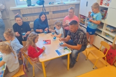 Grupa dzieci wraz z rodzicami siedzi przy prostokatnym stoliku i je niebieskie galaretki w przeźroczystych pucharkach . Na stoliku leży dyplom i kolorowa ksiażeczka . Przy półce stoi chłopiec w niebieskiej koszulce i spodenkach i je galaretkę.