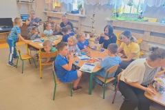 Sala przedszkolna . Rodzice i dzieci siedzą przy stolikach i jedzą niebieskie galaretki. Jeden chłopiec stoi przy stoliku.