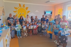 Sala przedszkolna . Dzieci i rodzice stoją w półkolu . Dzieci w strojach ekologicznych trzymają dyplomy . Z lewej strony widać szafki na półki indywidualne . W tle na ścianie namalowane żółte słoneczko i ekran telewizora.