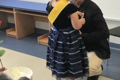 W sali przedszkolnej widać mężczyznę w okularach, ubranego w jasne spodnie, ciemną koszulę i czarną marynarkę. Przykucnął on przed dziewczynką, która ma na sobie ciemną elegancką sukienkę w błyszczące pasy, ciemne buty i białe podkolanówki. Przez ramię ma przełożoną żółtą szarfę. Przypina kartonowy medal do klapy marynarki mężczyzny. Z przodu widać fragment ramienia dziecka, które w dłoni ma biały talerzyk z mała szklaneczką soku. W tle niebieskie biurka i dekoracje ze słoneczek.