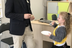 Z lewej strony stoi uśmiechnięty mężczyzna w okularach.  Ubrany jest w jasne spodnie, ciemną koszulę i czarną marynarkę. Trzyma on w dłoniach małą szklaneczkę z sokiem z cytryny. Podała mu go z białego talerzyka - tacy dziewczynka stojąca                z prawej strony. Ubrana jest w szary sweterek i ciemną spódniczkę. Dziewczynka ma uniesiony w górę kciuk. W tle widać biurko, szaro - zielone szafki oraz wiszący na ścianie telewizor.