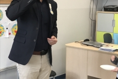 Mężczyzna w okularach ubrany w jasne spodnie, ciemną koszulę i czarną marynarkę przystawia do ust małą szklaneczkę z sokiem z cytryny. W tle dekoracja ścienna - symbol Orderu Uśmiechu : żółte słoneczko na okrągłym niebieskim tle oraz podobne emblematy wykonane na białych kartkach przez dzieci. Z prawej strony biurko i szafki.