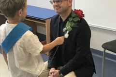 Mężczyzna w okularach ubrany w jasne spodnie, ciemną koszulę i czarną marynarkę przykucnął przed chłopcem ubranym w jasną koszulę i ciemne spodnie. Przez ramię chłopca przewieszona jest niebieska szarfa. Chłopiec przykłada czerwoną różę do prawego ramienia mężczyzny. W tle tablica z dekoracją oraz niebieskie biurko.