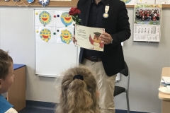 Mężczyzna w okularach  ubrany w jasne spodnie, ciemną koszulę i czarną marynarkę trzyma w ręku czerwoną różę oraz dyplom z wizerunkiem sowy  w akademickiej czapce. Napis jest nieczytelny. Zwrócony jest twarzą do dzieci, z których widać częściowo chłopca w białej koszuli i dziewczynkę w szarym sweterku. W tle dekoracja - Symbol Orderu Uśmiechu: żółte słońce na okrągłym niebieskim tle. Obok napis czarnymi drukowanymi literami na żółtym tle: Kawaler Orderu Uśmiechu. Na tablicach dekoracja emblematów orderu uśmiechu wykonanych przez dzieci.