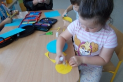 Troje dzieci siedzi przy stoliku przedszkolnym.  Przed dziewczynką ubraną w niebieską bluzkę           w białe kwiaty leży biała kartka z naklejonym niebieskim kołem, klej oraz małe kartoniki z niebieskimi serduszkami. Obok siedzi chłopiec  w granatowej koszulce z białym nadrukiem. Dzieci wycinają serduszka. Trzeci chłopiec w jasnej koszulce przygląda się pracy. Z tyłu widoczne drzwi wyjściowe z sali z naklejonymi niebieskimi sowami.