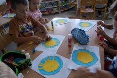 Przy stoliku przedszkolnym, po prawej stronie siedzi dziewczynka w jasnym ubraniu z nadrukiem pieska. Smaruje klejem żółte koło z kartonu. Obok leży wycięte większe koło w kolorze niebieskim. Wokół stolika siedzi jeszcze troje dzieci widocznych fregmentarycznie. Wycinają one koła z kartonu. Na stoliku leżą piórniki i nożyczki.