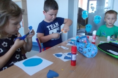 W sali przedszkolnej przy stoliku siedzi kilkoro dzieci, dwoje z nich jest dobrze widocznych. Przed nimi leżą białe kartki z naklejonymi mniejszymi żółtymi kołami na większe niebieskie. Chłopiec                          w brązowej koszulce z nadrukiem dokleja żółte promyczki do żółtego koła, podobnie robi siedząca obok niego dziewczynka w różowej bluzeczce. Pozostałe dzieci , które też to robią widoczne są fragmentarycznie. Na stoliku leżą piórniki i kleje.