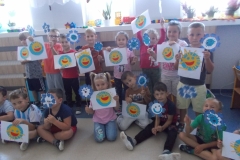 W sali przedszkolnej widoczna jest grupa dzieci . W pierwszym rzędzie z przodu siedzi siedmioro dzieci, za nimi w drugim rzędzie stoi ośmioro. Dzieci trzymają w rękach prace plastyczne: emblematy Orderów Uśmiechu oraz kwiaty z niebieskich serduszek na patyczkach. W tle widać okna.