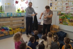 Na podłodze sali przedszkolnej siedzi odwrócona tyłem grupa dzieci. Patrzą           w kierunku ubranej w ciemne spodnie i szarą bluzkę nauczycielki. Trzyma ona w ręce otwarte pudełko. Obok stoi mężczyzna ( ojciec dziecka) ubrany w ciemne spodnie        i niebieską koszulę w kratkę. Trzyma on w dłoniach czarny materiał. Z lewej strony widać okna z wiosennymi dekoracjami, pod spodem stoją żółte pudełka z klockami. W głębi widoczna tablica tematyczna oraz powieszone obrazki z literami.