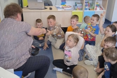 Grupa dzieci przedszkolnych siedzi w gromadce na podłodze. Z lewej strony siedzi na krzesełku nauczycielka, zwrócona jest twarzą do dzieci. Podaje coś jednemu           z chłopców. Dwie dziewczynki się zgłaszają podnosząc palce w górę. Kilkoro chłopców się uśmiecha. Za nimi widać biurko i kolorową szafkę na pomoce dydaktyczne.