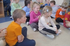 Dzieci siedzą w sali przedszkolnej na podłodze w trzech rzędach, jedno za drugim. Każde robi masażyk na plecach kolegi lub koleżanki. Po prawej stronie stoi biurko, na ścianie w głębi wisi biała tablica z planszą gospodarstwa.