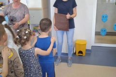 W sali przedszkolnej obok drzwi stoi młoda kobieta ( mama ) ubrana w jasno niebieskie spodnie i ciemną koszulkę. Trzyma w dłoniach poduszkę. Przed nią stoją ustawione rzędem dzieci. Z lewej strony stoi nauczycielka mówiąca coś do dzieci. Za nią znajdują się biało - szare szafki. Obok drzwi stoi żółty kosz na śmieci.