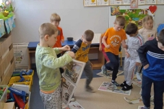 Dzieci przedszkolne ubrane w kolorowe stroje depczą po arkuszach gazety. Jeden z chłopców stojących na przodzie trzyma gazetę w rękach. W tle widoczna tablica tematyczna i tablica z literami oraz szafki na pomoce dydaktyczne.