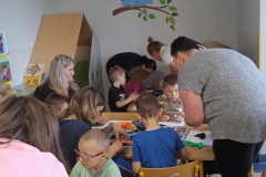 Przy stolikach przedszkolnych siedzą dzieci. Obok siedzą lub stoją mamy, pochylając się nad stolikami. Tworzą prace plastyczne. Na stole widać kartki papieru i bibuły. Na ścianie w głębi widać namalowaną siedzącą na gałęzi sowę. Jest ona w okularach i czyta niebieską książkę.      Z lewej strony stoi szafka z książkami i kącik wypoczynkowy.