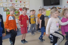 Dzieci ustawione w gromadzie wykonują ćwiczenia ruchowe. Kilkoro stoi na jednej nodze, niektórzy lekko ją unoszą w górę. Z prawej strony na ścianie wisi czarny telewizor. Za dziećmi znajduje się tablica tematyczna oraz przypięte u góry na tablicy plansze z literami.