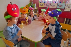 Starszaki siedzą przy stoliku, na głowach mają kolorowe opaski z owocami, jedzą przygotowane przez siebie szaszłyki owocowe.