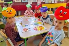 Dzieci siedzą przy stoliku, na głowach mają kolorowe opaski z owocami, wydzierają owoce z papieru kolorowego.