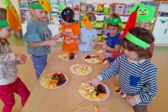 Dzieci stoją wokół stołu  na głowach mają kolorowe opaski z owocami, na stole leżą talerze z pokrojonymi owocami, dzieci nabijają na patyczki, robią szaszłyki.