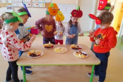 Kilkoro dzieci stoi wokół stolika na którym leżą talerze z pokrojonymi owocami. Nabijają je na patyczki, robią owocowe szaszłyki. Na głowach mają opaski z kolorowymi owocami.