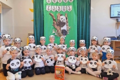 Grupa dzieci siedzi na beżowej podłodze. W rękach trzymają głowę pandy z kartonu. Na głowach dzieci opaski pandy z papieru. W tle na zielonym płótnie napis: Dzień Pandy. Pod nim biało- czarno panda na brązowym drzewie.