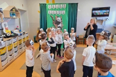 Dzieci siedzą w rzędzie przy szafkach. Na środku sali leży poducha, skakanka, niebieska i żółta obręcz. Dziewczynka w czarnych spodniach i białej bluzce pokonuje tor przeszkód.