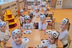 Dzieci siedzą na krzesłach przy 4 stolikach. Na głowie mają maski pandy. Przed nimi na stole biało- czarne głowy pandy z kartonu. W tle regały z zabawkami.