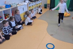 Dzieci siedzą w rzędzie przy szafkach. Na środku sali leży poducha, skakanka, niebieska i żółta obręcz. Dziewczynka w czarnych spodniach i białej bluzce pokonuje tor przeszkód.