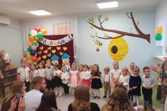 Dzieci ustawione w półkolu stoją przed rodzicami. W tle na bordowym płótnie napis "Pasowanie". Pod napisem kolorowe serduszka z imionami dzieci. Z boku zawieszone balony.