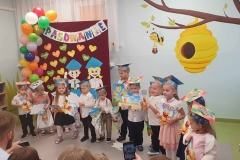 Grupa dzieci w kolorowych czapkach stoi w półkolu . W rękach trzymają dyplomy i worki z prezentami.