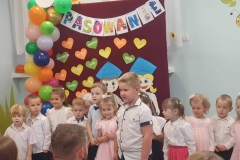 Chłopiec w białej koszuli stoi przed grupą dzieci. Za nim w tle kolorowa dekoracja.