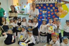 Pięcioro dzieci siedzi na podłodze,  pozostała grupa oraz dwie  panie stoją za nimi. W tle dekoracja. Na niebieskim tle widnieje  napis Pasowanie, są  też  kolorowe liście.