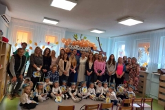 Grupa dzieci ubranych w białe koszule i bluzki,  ciemne spodnie  i spódniczki siedzi na podłodze. Na głowach każde z nich ma kolorową czapkę.  Za nimi  stoją panie i pan.  W tle widać pomarańczowe balony,  niebieską  ścianę i okna.