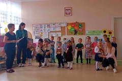 Grupa dzieci stoi w dwóch rzędach, w pierwszym Dzieci pasowane, na głowach mają kolorowe czapki, na szyi zawieszone głowy Misie. Z lewej strony widać dwie panie. W tle kolorowe dekoracje.